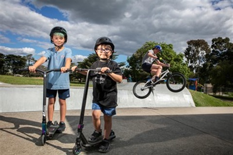 Three children are at the Whittlesea Skate Park. Two hold their scooters and look at the camera, and another one rides the ramp in the background on their bike