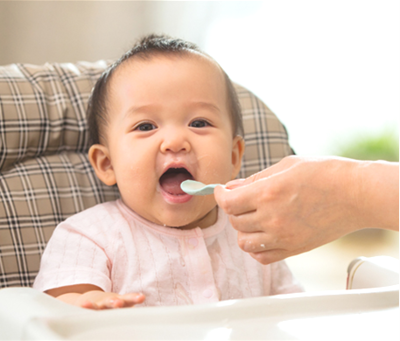 A baby sits in a highchair. An adult hand spoons food into the child's mouth.